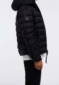 Zwarte pufferjas met capuchon, voorzien van een ritssluiting en zijzakken. Gestructureerde stof met een opvallend merklogo op de mouw.