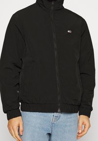 Černá bunda s vysokým límcem, vybavená předním zipem a elastickými manžetami. Hladká tkanina s minimalistickým logem na hrudi.