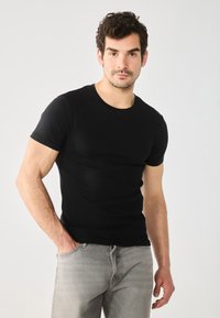 Schwarzes Kurzarm-T-Shirt mit Rundhalsausschnitt, figurnaher Schnitt mit glatter Textur, getragen mit hellgrauen Jeans, Hände in den Taschen, neutraler Hintergrund.