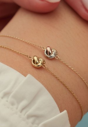Bracelets en double chaîne en or et en argent avec des breloques en forme de nœud interconnectées. Finition métallique lisse, design minimaliste et texture délicate.