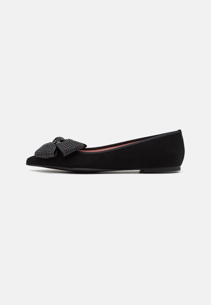 Ballerine noire avec un nœud texturé sur le bout et un petit talon, sur un fond clair uni.