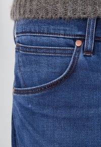 Vaqueros de denim azul con una textura suave, que presentan costuras naranjas, un botón de tono cobre y un detalle en el bolsillo delantero.