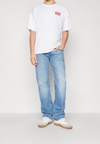 Vit kortärmad t-shirt med en röd Diesel-logotyp på bröstet, ihop med ljusblåjeans och vita sneakers med beige detaljer.