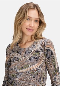 Frau mit schulterlangem blonden Haar, die ein langärmliges Oberteil mit Paisley-Muster in beige, schwarz, grün und orange trägt und sanft lächelt.
