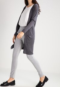 Cardigan lungo aperto di colore grigio scuro, realizzato in materiale leggero. Abbinato a pantaloni grigi chiari a gamba affusolata e mocassini neri.