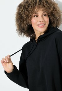 Černá mikina s kapucí, stahovací šňůrkou a volným střihem. Měkká textura a dlouhé rukávy. Minimalistický design bez viditelných vzorů nebo akcentů.
