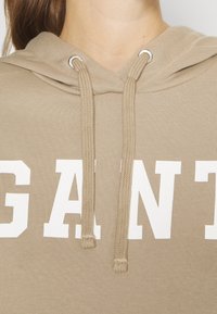 Béžová mikina s kapucí, která má šňůrku se stříbrnými konci, velký bílý nápis "GANT" a měkkou, strukturovanou látku.