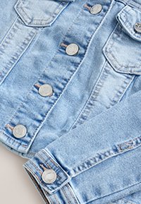 Hellblaue Denimjacke mit silbernen Metallknöpfen und sichtbaren Nähten an den Brusttaschen und dem Ärmelsaum.