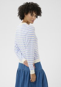 Femme aux cheveux bouclés jette un coup d'œil par-dessus son épaule, portant un pull crème à rayures bleu clair et une jupe en denim bleu, sur un fond blanc.