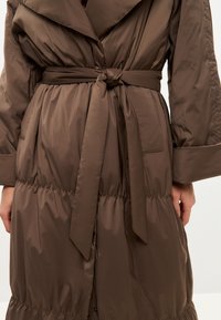 Cappotto imbottito marrone con vita cinturata, dotato di un ampio colletto, tasche laterali e una texture trapuntata.