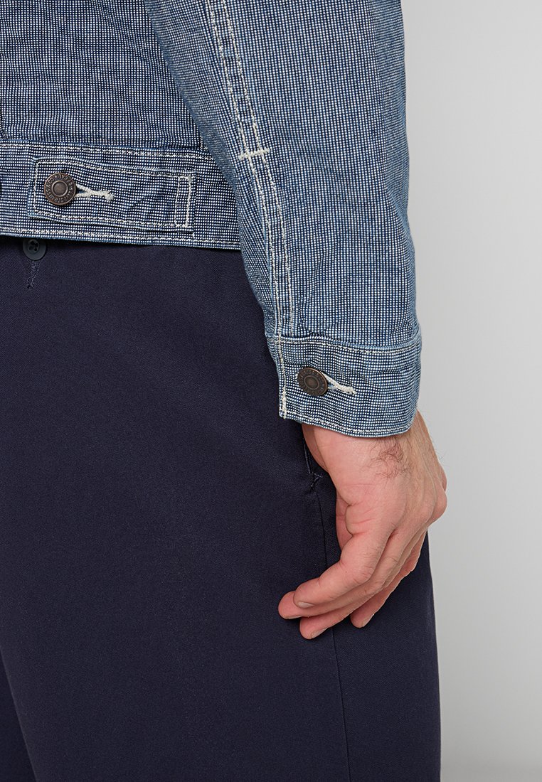 Veste en jean avec un motif à carreaux, de couleur bleue, et des boutons marron contrastés aux poignets, associée à un pantalon bleu marine.
