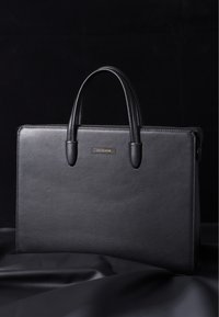 Sort læderportefeuille med tekstureret overflade, to top håndtag og et sølv logo på forsiden. Glat lynlåslukning på toppen.