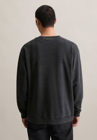 Man met kort zwart haar in een simpele zwarte longsleeve sweatshirt, gezien van achteren tegen een neutrale achtergrond.