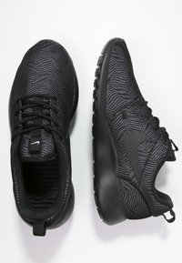 Svarta sportskor i strukturerad tyg med fiskbensmönster, med en slät svart sula och minimalistisk märkning på tungan.