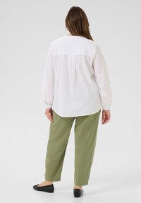 Femme se tenant de dos, portant une chemise blanche à manches longues, un pantalon vert olive et des chaussures plates noires devant un fond uni.