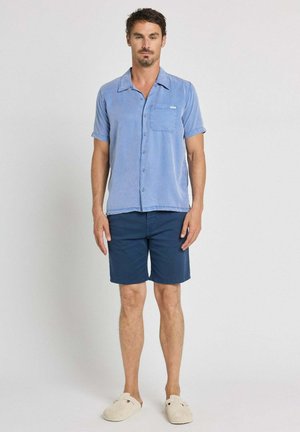 Homme debout portant une chemise bleu clair à manches courtes avec boutons, un short bleu foncé et des chaussures beige à enfiler sur un fond uni.