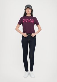 Violetinė marškinėliai su rožiniu "VERSACE JEANS COUTURE" logotipu, derinami su tamsiai mėlynomis liesomis džinsomis ir baltais sportbačiais. Sidabriniai akcentai ant kepurės.
