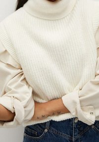 Pull à col roulé blanc côtelé superposé à un chemisier crème à manches bouffantes, porté avec un jean en denim bleu. Tatouage visible sur le poignet.