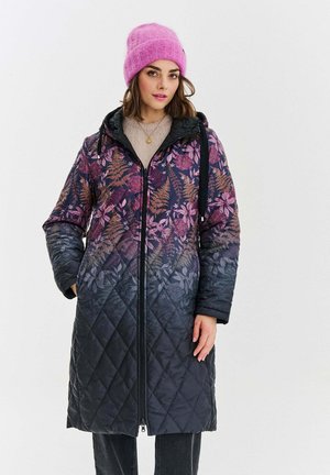 Kobieta nosząca długi pikowany płaszcz w kwiatowe wzory w odcieniach purpury i czerni, w zestawieniu z różowym dzianinowym czapką i czarnymi spodniami.