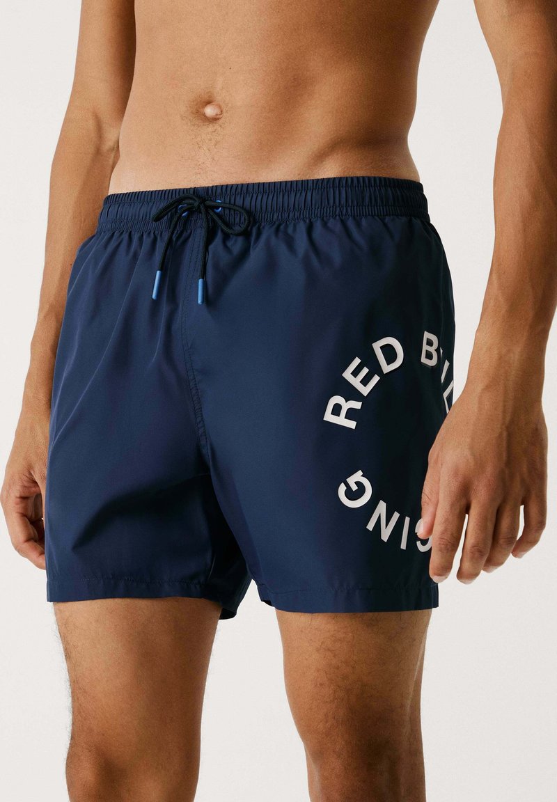 Pantaloni da bagno blu navy con vita elasticizzata, cordoncino regolabile e testo bianco "RED BULL" stampato sul lato. Texture liscia.