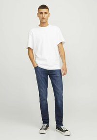 Camiseta de algodón blanca de manga corta, vaqueros de denim azul y zapatillas negras con suelas blancas, en una pose de pie frente a un fondo neutro.