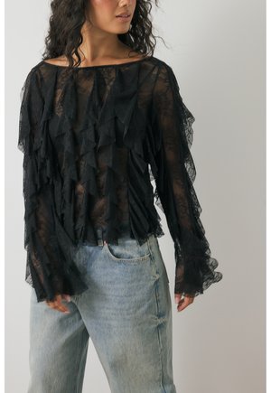 Woman wearing a black sheer lace top with layered ruffles and light blue high-waisted jeans, standing against a plain background.