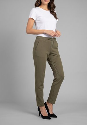 Mujer con camiseta blanca de manga corta, pantalones verde oliva con cordón y bolsillos laterales, y zapatos de tacón negros de pie sobre un fondo gris.