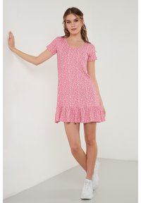 Korte roze jurk met een uitlopende zoom, versierd met een wit bloemetjespatroon en korte mouwen. Draagt met witte sneakers.