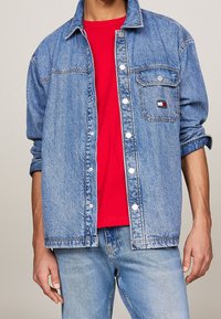 Man wearing light blue denim jacket over red t-shirt and blue jeans, jacket pocket features a small Tommy Jeans logo patch.