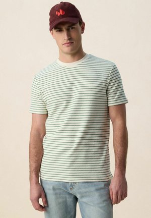 Homme portant une chemise blanche à manches courtes avec de fines rayures horizontales vertes, un jean bleu clair et une casquette bordeaux avec un logo orange.