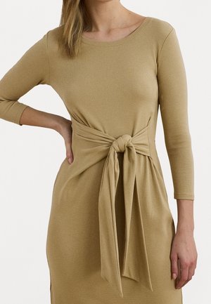 Kvinde iført en beige langærmet kjole med et stort knyttet bælte ved taljen, stående med den ene hånd på hoften.