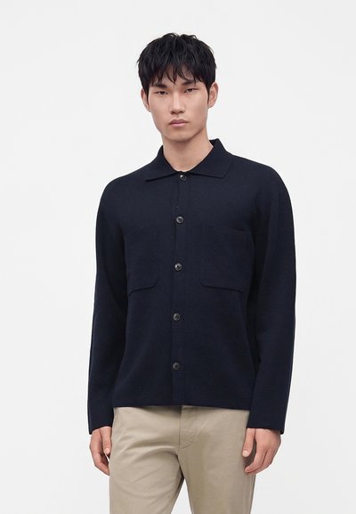 Jeune homme portant un cardigan noir boutonné avec deux poches poitrine et un pantalon beige, debout devant un fond blanc uni.