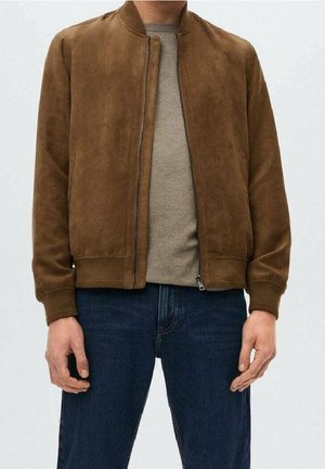 Mężczyzna ubrany w brązową zamszową kurtkę bomberkę na beżowym swetrze i ciemnoniebieskich dżinsach, stojący z rękami swobodnie opuszczonymi wzdłuż ciała.