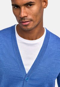 Blauer Cardigan mit V-Ausschnitt und strukturiertem Strick. Verfügt über einen einzelnen blauen Knopf an der Vorderseite, getragen über einem weißen T-Shirt.