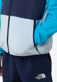 Világoskék és tengerészkék polárdzseki cipzárral, oldalzsebbel és logóval sötét nadrágon. Puha tapintású, színes blokkokból álló dizájn.