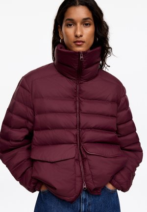 Femme aux longs cheveux foncés portant une veste matelassée bordeaux zippée avec un col montant et des poches, debout les mains dans les poches de la veste.