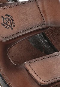 Sandalia de cuero marrón con correas de Velcro, detalle de logo circular y una plantilla texturizada que muestra un diseño minimalista y bordes suaves.