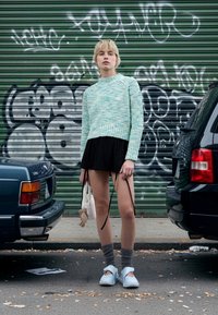 Ανοιχτό μπλε κοντό πουλόβερ με υφή πλέξης, μαύρη φούστα με πλαϊνές κορδέλες, γκρι γόνατο κάλτσες και ανοιχτά μπλε παπούτσια. Αστικό περιβάλλον με γκράφιτι.