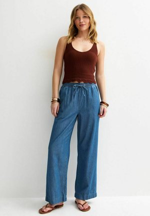 Vrouw staat tegen een muur, draagt een bruin gebreid mouwloos topje, losse blauwe spijkerbroek, bruine sandalen en meerdere armbanden.