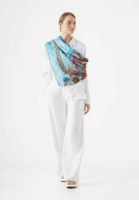 Écharpe en soie bleu vif avec des motifs floraux et géométriques, drapée sur une blouse blanche, avec des accents d'orange et de marron.