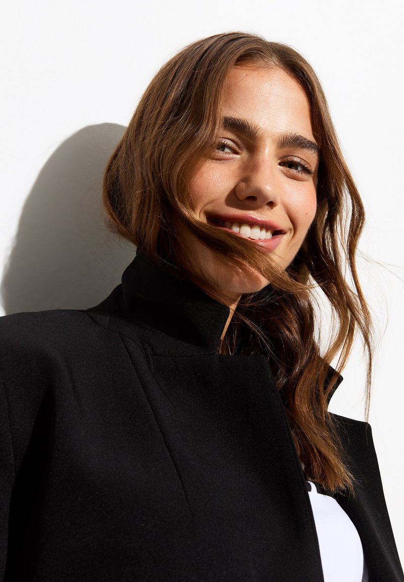 Veste noire à col montant avec des épaules structurées, texture lisse et design sur mesure, complétée par des cheveux naturels et en désordre.