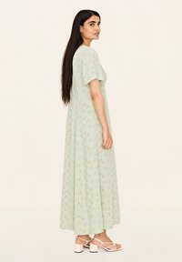 Hellgrünes Maxikleid mit Blumenmuster, kurzen Ärmeln, lockerer Passform und fließender Textur; kombiniert mit weißen Riemchensandalen.