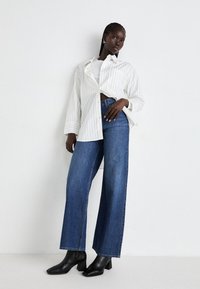 Donna in piedi contro un muro bianco, con una camicia bianca a righe sottili sopra una maglietta bianca, jeans blu a gamba larga e stivaletti neri con tacco.