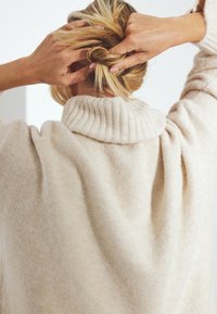 Beiger Strickpullover mit einem gerippten Kragen, der eine weiche Textur und eine lockere Passform aufweist. Die Person stylt ihre Haare mit den Händen.