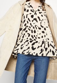 Femme portant un manteau en peau lainée beige sur un chemisier crème avec des taches abstraites noires et un jean bleu, cadré de la poitrine aux cuisses.
