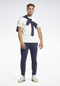 Hvid t-shirt med logo, marineblå sweatpants, marineblå sweater båret om skuldrene, og grå sportsko med farverige accenter.