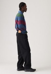 Maglietta a maniche lunghe a righe nei colori blu, verde e bordeaux; abbinata a pantaloni di jeans neri; etichetta visibile sulla tasca posteriore; calzature informali.