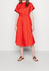 Kvinna klädd i en ljus röd midiklänning med korta ärmar, en krage och ett midjeband, tillsammans med bruna platta sandaler.