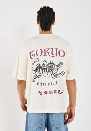 Osoba z warkoczami, nosząca białą koszulkę z czarnym tygrysem i napisem "Tokyo Shinjuku" oraz japońskimi znakami na plecach.