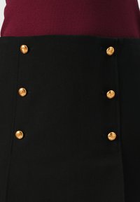 Jupe noire avec six boutons dorés sur le devant, au tissu lisse et à la coupe ajustée, associée à un haut bordeaux.